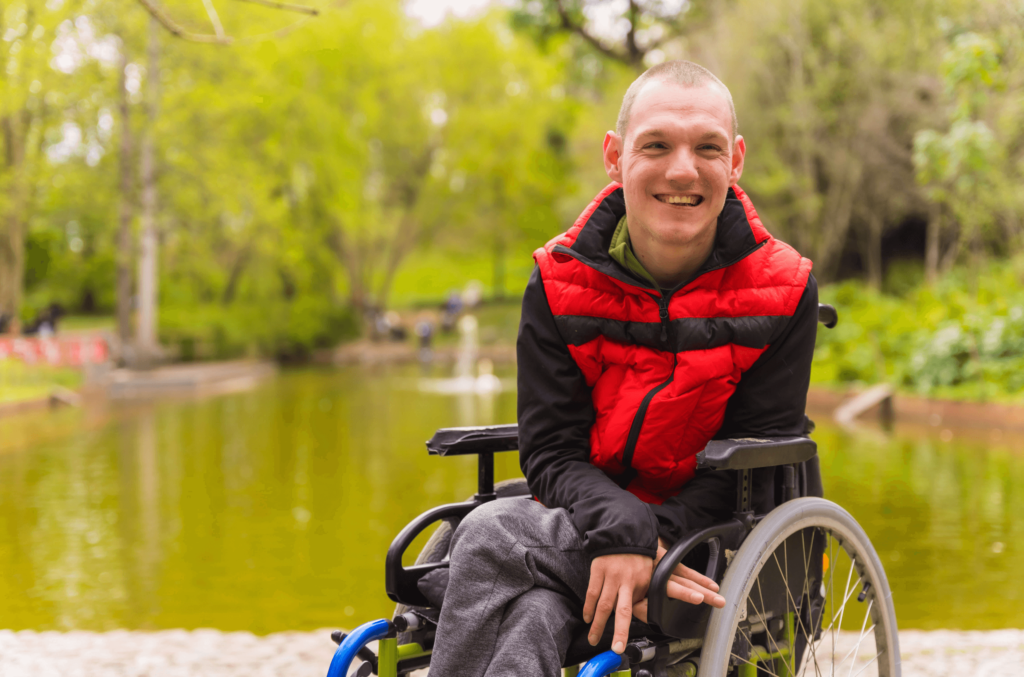 A young man in a wheelchair, wearing a red and black jacket, smiles brightly in a lush park setting, representing the freedom and security a first party special needs trust can provide.