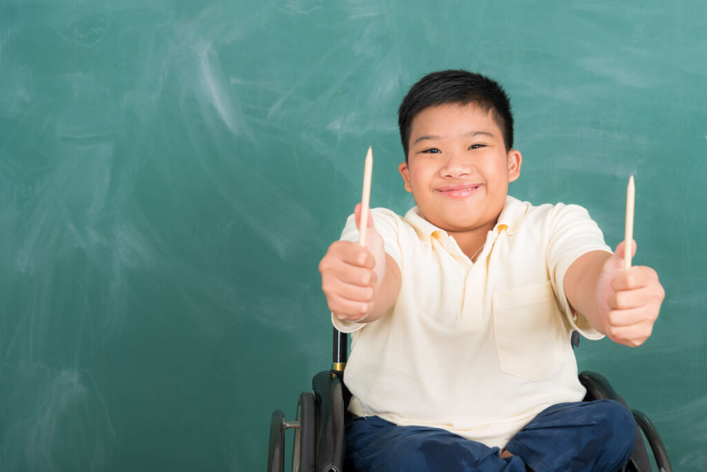 In a classroom setting, a young boy with a beaming smile is seated in a wheelchair, wearing a pale yellow polo shirt and navy pants. He holds a pencil in each hand, with thumbs up, symbolizing enthusiasm and a proactive stance towards learning. The green chalkboard in the background serves as a reminder of the educational journey ahead, where legal tools like the power of attorney for education ensure that his special needs are acknowledged and accommodated, allowing him to thrive academically.
