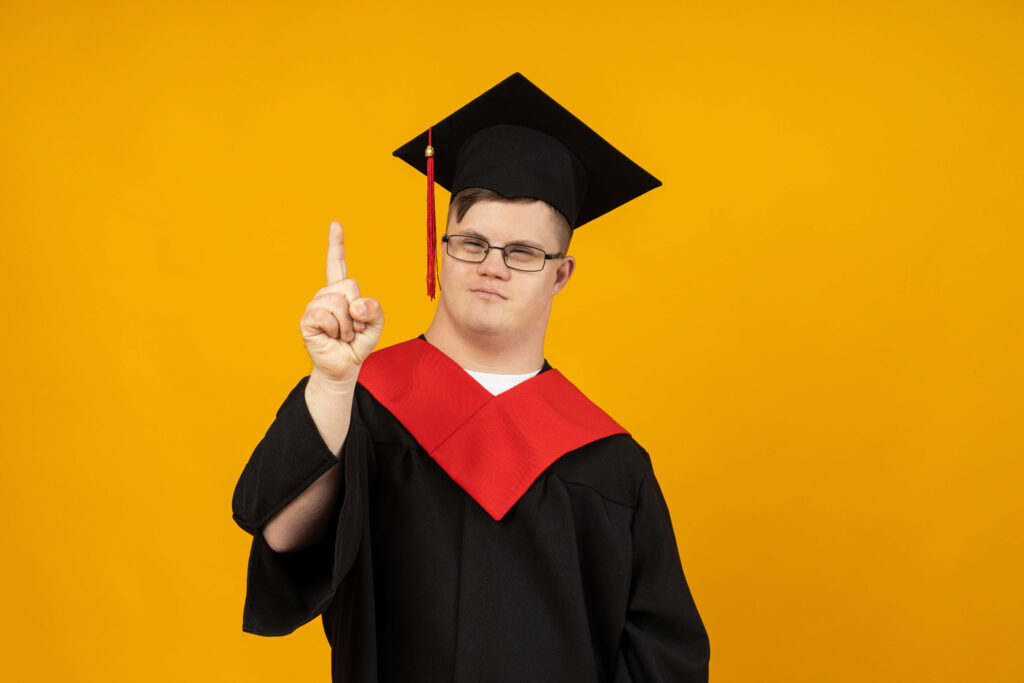 A proud graduate with Down syndrome, wearing a black gown and red stole, points upward against a yellow backdrop, signifying achievement and the role of power of attorney for education in his journey.