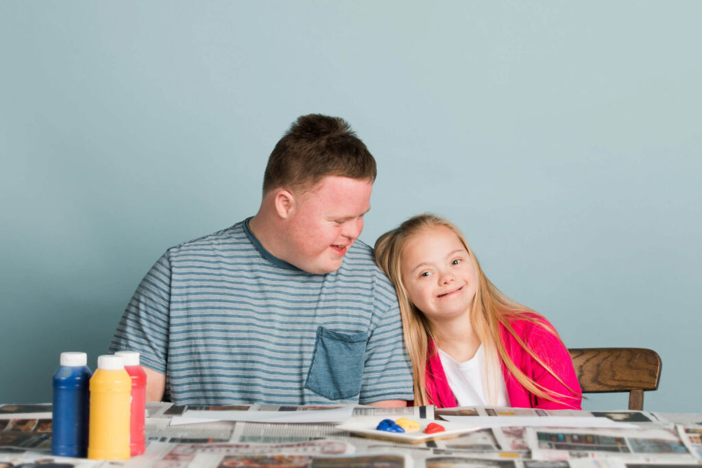 A young man and a girl with Down syndrome sit side by side, smiling and enjoying a painting activity, exemplifying the nurturing relationships supported by a power of attorney.
