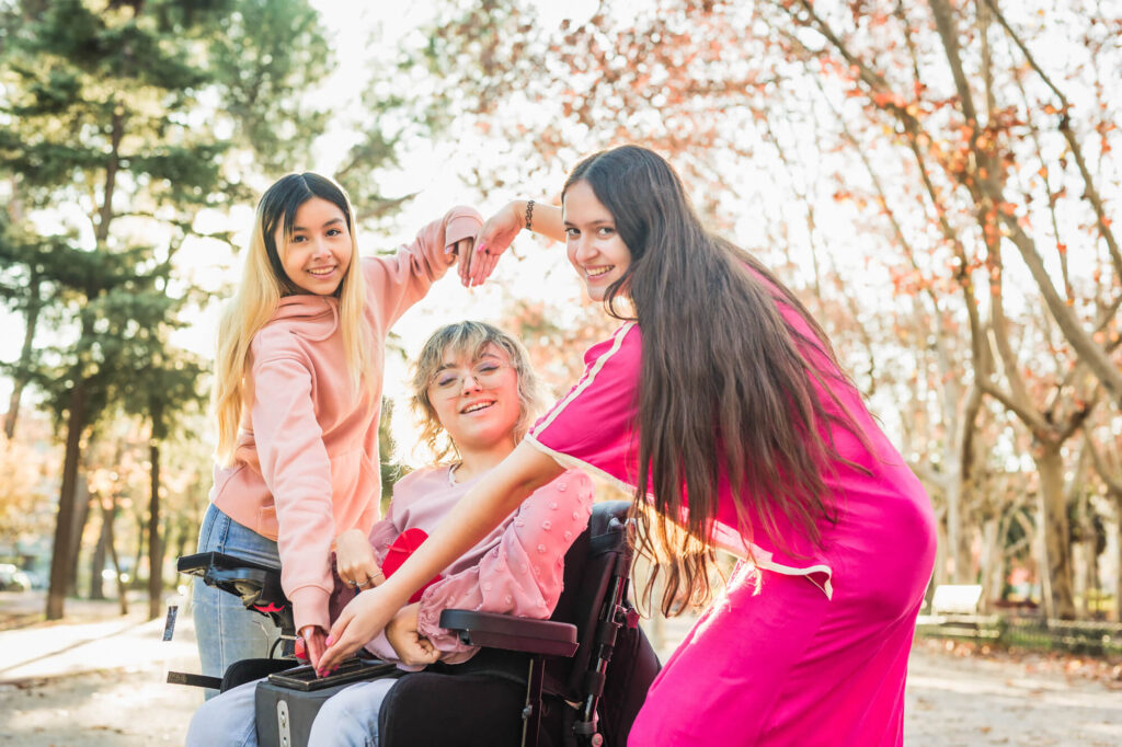 Three happy young women in a park, one in a wheelchair, enjoy a sunny day, highlighting the supportive community aspect of special needs trusts.