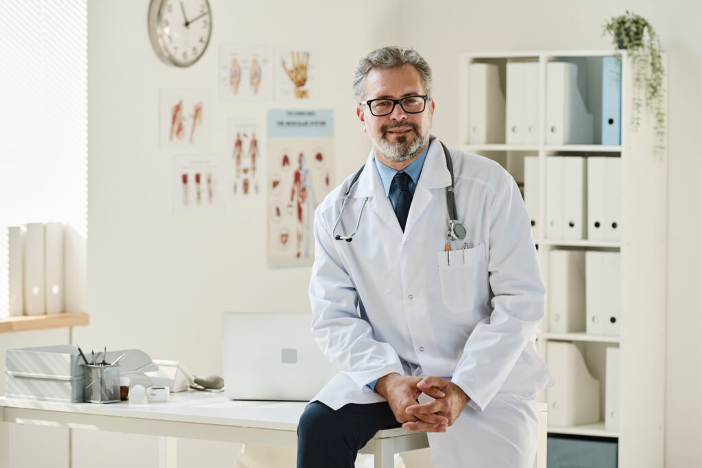 A confident, middle-aged doctor in a white coat with a stethoscope around his neck sits on a desk in a clinic, representing the importance of discussing healthcare decisions and power of attorney with medical professionals.